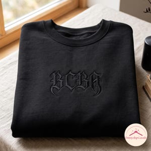 Pode incluir: Uma camisola preta de gola redonda com as letras BCBA bordadas em fio preto a condizer no centro do peito. O texto é apresentado num tipo de letra de inspiração gótica com extremidades afiadas e detalhes decorativos complexos.