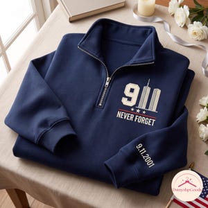May include: A navy blue quarter-zip sweatshirt featuring a graphic on the chest with the number 9, an illustration of the Twin Towers, three stars, and the text NEVER FORGET. The left sleeve is embroidered with the date 9.11.2001 in white thread.