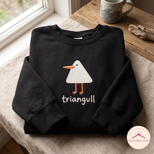 May include: A black long-sleeve sweatshirt featuring an embroidered white triangle-shaped bird with an orange beak and legs. Below the bird, the text "triangull" is embroidered in white thread. The garment has a crew neckline and ribbed cuffs.