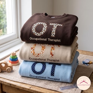 May include: A stack of three crewneck sweatshirts in dark brown, beige, and light blue. Each features the letters OT in a patterned fabric appliqué and the text Occupational Therapist embroidered below in a matching thread color.