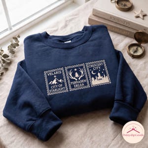 May include: A navy blue crewneck sweatshirt featuring three rectangular stamp-style embroidered designs in white thread. The designs depict mountain peaks labelled Velaris City of Starlight, deer antlers labelled Terrasen Erilea, and a city skyline labelled Crescent City.