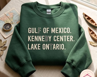 May include: A dark green crewneck sweatshirt featuring cream-colored text that reads GULF OF MEXICO. KENNEDY CENTER. LAKE ONTARIO. The letters F, D, and T are highlighted with a distinct outline style within the printed text on the front.