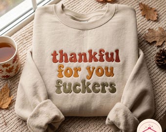 May include: A beige crewneck sweatshirt featuring embroidered text in a retro font. The word thankful is in burnt orange, for you is in golden yellow, and fuckers is in olive green. The sweatshirt has long sleeves and a ribbed collar.