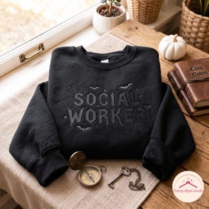 May include: A black crewneck sweatshirt featuring the embroidered text SOCIAL WORKER in a gothic font, surrounded by spider web and bat motifs. The design is centred on the chest of the long-sleeved garment, which is made of a solid black fabric.