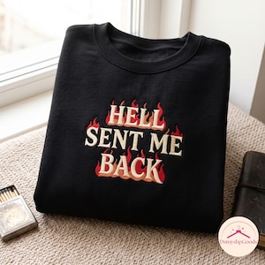 以下が含まれることがあります： クリーム色の文字で「HELL SENT ME BACK」と刺繍された黒いクルーネックのスウェットシャツ。文字の周りには赤とオレンジの炎の刺繍が施されており、衣服の前面に大胆なグラフィックデザインが描かれています。