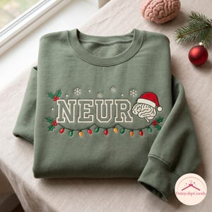 Könnte beinhalten: Ein olivgrünes Sweatshirt mit Rundhalsausschnitt und dem gestickten Text NEUR in College-Schrift. Rechts trägt eine Gehirn-Illustration eine rote Weihnachtsmütze. Das Design zeigt Stechpalmenblätter, rote Beeren, Schneeflocken und Lichterketten.