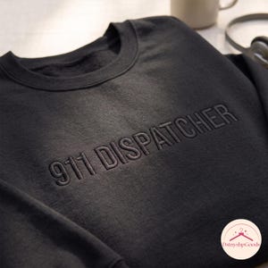 Könnte beinhalten: Ein schwarzer Rundhals-Pullover mit dem Text 911 DISPATCHER, der in passendem schwarzem Garn auf der Brust eingestickt ist. Das Kleidungsstück hat einen gerippten Halsausschnitt und lange Ärmel und zeigt ein monochromes Design mit sauberer Textur.