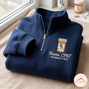 May include: A navy blue quarter-zip sweatshirt featuring an embroidered orange prescription bottle with white pills and the text Naomi, CPhT PHARMACY TECH. The garment has a high collar, a silver-toned metal zipper, and ribbed cuffs.