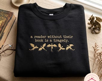 May include: A black crewneck sweatshirt featuring gold embroidery that reads, "a reader without their book is a tragedy." Below the text are five distinct gold embroidered dragon silhouettes arranged in a horizontal row across the chest.