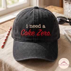 Op de afbeelding: Een zwarte baseballpet met een verweerde, gewassen textuur. De voorkant is voorzien van geborduurde tekst in twee regels: de woorden "i need a" in het wit, gevolgd door de woorden "Coke Zero." in een vetgedrukt, rood cursief lettertype.