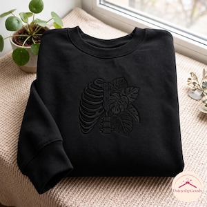 May include: A black crewneck sweatshirt featuring a tonal black embroidered design on the chest. The embroidery depicts a human rib cage on the left side, seamlessly integrated with detailed monstera leaves on the right side.