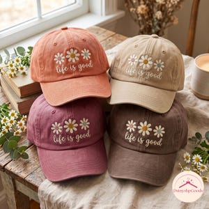 Puede incluir: Cuatro gorras de béisbol en tonos coral, beige, granate y marrón oscuro. Cada gorra presenta un diseño bordado de tres margaritas y el texto "life is good" en letras cursivas blancas en el panel frontal, justo encima de la visera curva.