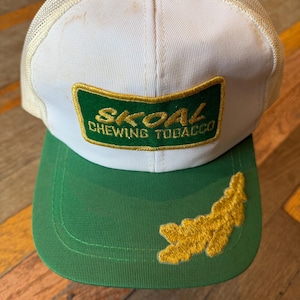 May include: A white and green trucker hat featuring a rectangular patch with the text SKOAL CHEWING TOBACCO in gold embroidery. The hat has a white mesh back, a green brim, and a gold embroidered leaf-shaped design on the right side of the brim.
