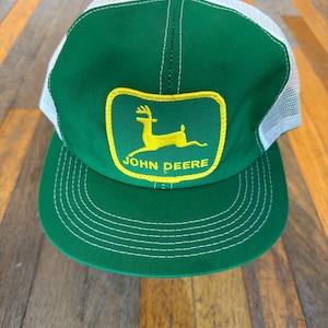 May include: A green and white trucker hat featuring a yellow patch on the front panel. The patch displays a yellow leaping deer silhouette above the text JOHN DEERE. The hat has a solid green front, a white mesh back, and a green brim with stitching.