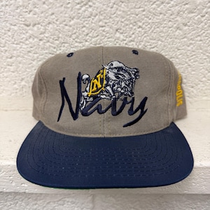 Puede incluir: Una gorra de béisbol color topo con visera azul oscuro. La parte frontal presenta la palabra Navy bordada en azul marino y un gráfico de una mascota cabra con una N amarilla. El lateral de la gorra tiene la palabra Annapolis bordada en hilo amarillo.