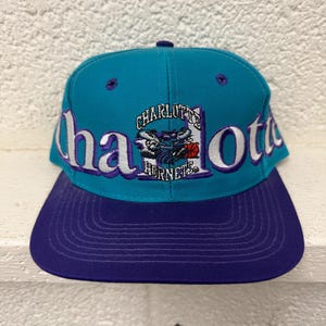 Könnte beinhalten: Eine türkisfarbene Baseballkappe mit violettem Schirm, die vorne mit einem gestickten Charlotte Hornets-Logo und einer Basketball-Grafik versehen ist. Das Wort Charlotte ist in großen weißen und violetten Buchstaben auf die Vorderseite gestickt.