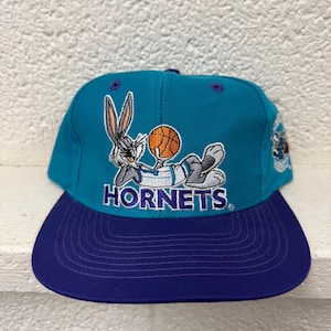 May include: A teal and purple snapback cap featuring an embroidered graphic of Bugs Bunny holding a basketball above the word HORNETS in purple lettering. The cap has a structured crown with purple eyelets and a solid purple flat brim.