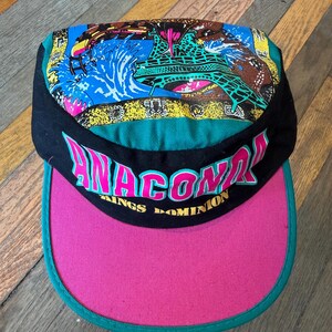 Könnte beinhalten: Eine schwarze Kappe mit pinkfarbenem Schirm und türkisfarbenem Rand. Vorne steht der Text ANACONDA in Pink und KINGS DOMINION in Gelb. Das Oberteil zeigt eine bunte Grafik einer Achterbahn, Wasserspritzern und einem Schlangenkopf auf blauem Grund.