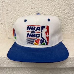 Op de afbeelding: Een witte honkbalpet met een blauwe klep en een geborduurd NBA on NBC-logo op de voorkant. Het logo bevat de tekst NBA ON NBC, een wit silhouet van een basketballer en een kleurrijk pauwenwaaierontwerp aan de onderkant.