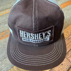 May include: A brown baseball cap with a mesh back and a rectangular patch on the front. The patch features the white text HERSHEY'S MILK CHOCOLATE. The cap has white stitching details along the brim and the crown, with a structured front panel.