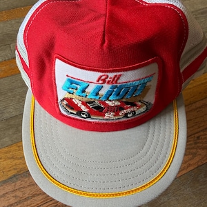 May include: A red and white trucker hat featuring a rectangular patch on the front. The patch displays the text Bill Elliott in blue letters above an illustration of a red race car with the number 9. The hat has a grey brim with yellow trim.