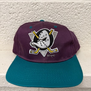 Può includere: Un cappellino da baseball viola con visiera color verde acqua che presenta un logo ricamato di una maschera da portiere a forma di papero bianco sopra due bastoni da hockey gialli incrociati su sfondo triangolare nero e argento con Disney.