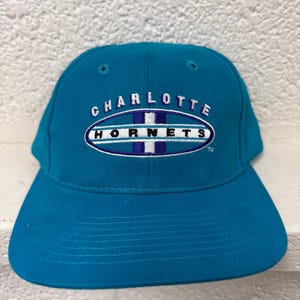 Op de afbeelding: Een talingkleurige honkbalpet met een geborduurd Charlotte Hornets-logo op de voorkant. Het logo bestaat uit het woord CHARLOTTE in witte letters boven een ovale vorm met het woord HORNETS in zwarte letters op een wit en paars gestreepte achtergrond.