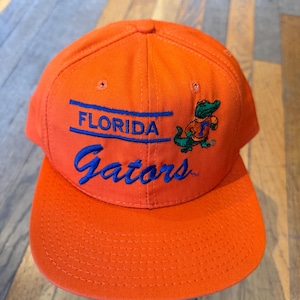 May include: A bright orange baseball cap featuring the embroidered text FLORIDA in blue block letters above the blue script word Gators. To the right of the text is an embroidered green alligator mascot wearing an orange shirt with a blue F.