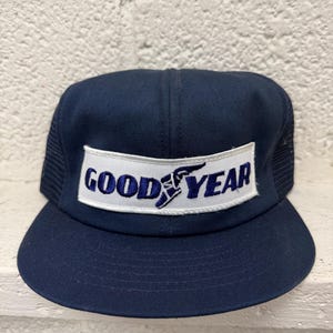 Könnte beinhalten: Eine marineblaue Baseballkappe mit einem weißen rechteckigen Aufnäher auf der Vorderseite. Der Aufnäher zeigt den Schriftzug GOODYEAR in dunkelblauen Buchstaben, wobei das ikonische Flügel-Logo in die Mitte integriert ist.