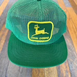 May include: A green mesh trucker hat featuring a yellow rectangular patch on the front. The patch displays a yellow leaping deer silhouette above the text JOHN DEERE in yellow capital letters. The hat has a solid green brim and a button on top.