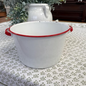 Peut inclure: Une bassine en émail blanc avec une finition lisse et un bord rouge vif. Le bol rond présente deux petites poignées rouges assorties sur les côtés opposés. La surface montre des signes d'usure mineurs, dont de petites taches sombres.