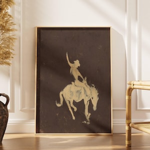 Può includere: Una stampa artistica da parete rettangolare incorniciata con uno sfondo marrone scuro e una silhouette color beige chiaro, in stile pittorico, di un cowboy che cavalca un cavallo imbizzarrito. La figura è ritratta di profilo con un braccio alzato.