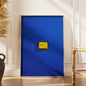 May include: A rectangular wall art print featuring a solid deep blue background with a single, centered yellow computer keyboard key. The key is square-shaped with rounded corners and has the word Enter printed in black on its surface.