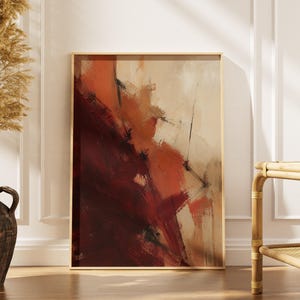 May include: A rectangular framed wall art piece featuring an abstract composition with bold, sweeping brushstrokes in deep burgundy, burnt orange, and cream tones, set within a thin, light-colored wooden frame.