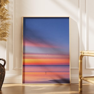 Puede incluir: Una impresión de arte mural rectangular enmarcada que muestra una puesta de sol sobre un océano en calma. El cielo pasa de un azul profundo en la parte superior a tonos vibrantes de naranja y rosa cerca del horizonte. Una pequeña silueta oscura.