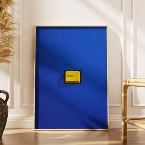 May include: A rectangular wall art print featuring a solid deep blue background with a single, centred yellow computer keyboard key. The key is square-shaped with rounded corners and has the word Enter printed in black on its surface.