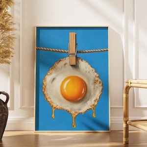May include: A framed wall art print featuring a fried egg with a bright orange yolk hanging from a horizontal rope by a wooden clothespin against a solid blue background. The egg white has golden liquid dripping from the bottom edge.