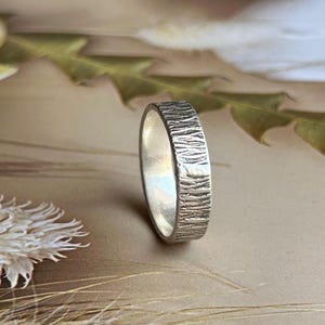May include: A silver ring with a textured, bark-like finish on the outer surface. The band has a consistent width and a metallic sheen, featuring vertical grooves and ridges that create a rugged, organic appearance across the entire exterior.