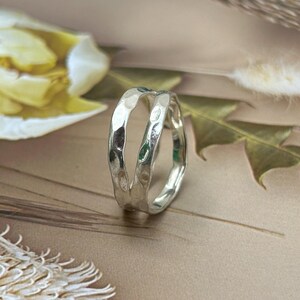 Può includere: Un anello in argento con design a doppia fascia e finitura martellata e testurizzata. Le due fasce sono collegate alla base, creando uno spazio aperto tra loro. La superficie metallica riflette la luce, evidenziando il motivo irregolare.