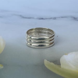May include: A polished silver ring featuring a triple-band design with three distinct, parallel horizontal grooves circling the entire circumference of the metal band, creating a stacked appearance with a smooth, reflective finish.