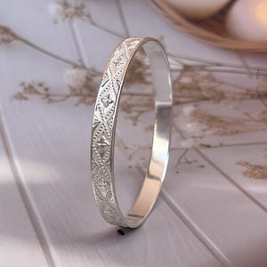 Puede incluir: Una pulsera rígida de metal en tono plateado que presenta un patrón repetitivo de diamantes en relieve con pequeños motivos de estrellas de cuatro puntas centrados en cada forma. La superficie exterior tiene un acabado texturizado con bordes pulidos.