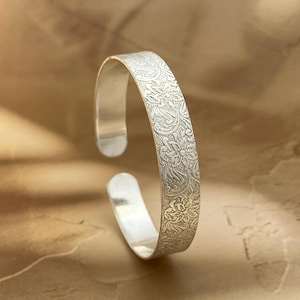 May include: A silver-toned cuff bracelet featuring an intricate, embossed floral pattern across its outer surface. The bracelet has an open-ended design with smooth, polished inner edges and a consistent width throughout its circular structure.
