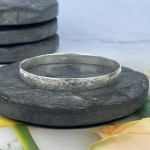 May include: A silver bangle bracelet with a hammered, textured surface. The circular metal band features a reflective finish and a consistent width throughout its design, showcasing a metallic sheen that catches the light across its uneven surface.