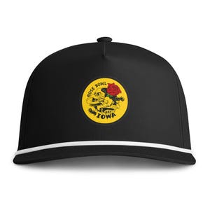 Puede incluir: Una gorra de béisbol negra con un cordón blanco en la base de la corona. La parte frontal presenta un parche circular amarillo con un gráfico negro de una mascota de pájaro, una rosa roja y el texto ROSE BOWL 1957 IOWA en letras negras.