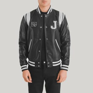 Pode incluir: Uma jaqueta varsity preta com mangas de couro sintético branco e acabamentos canelados listrados. A frente apresenta um emblema com a letra J branca no peito esquerdo e um emblema retangular com o texto EST. 1787. Possui botões de pressão prateados.