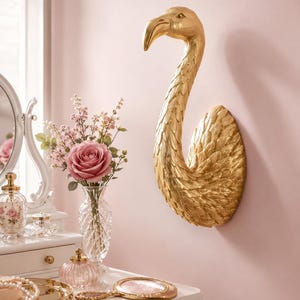 Può includere: Una scultura da parete color oro raffigurante la testa e il collo di un fenicottero con dettagliate texture di piume. L'oggetto presenta un collo curvo e un becco prominente, rifinito con una lucentezza metallica che riflette la luce.