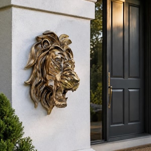 May include: A three-dimensional wall sculpture of a lion head finished in a metallic bronze tone. The lion is depicted with a full, flowing mane and an open mouth, showing teeth in a roaring expression. The piece is mounted on a flat vertical surface.