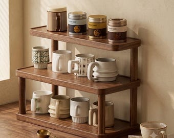 May include: A two-tiered wooden display shelf holding four cylindrical metal canisters on the top shelf and seven ceramic mugs of various shapes and textures on the middle and bottom shelves, with a small gold-toned metal spoon resting nearby.