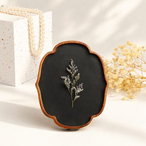 May include: A scalloped wooden frame with a dark matte interior surface featuring a single pressed sprig of dried purple flowers. The frame has a warm brown wood grain finish and a distinct quatrefoil shape, displaying a botanical specimen.