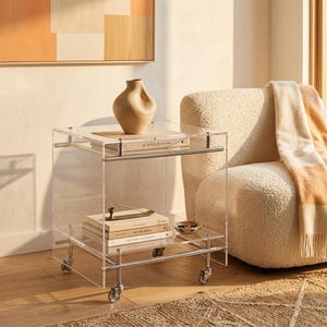 May include: A clear acrylic rolling side table with two shelves and silver metal hardware. The top shelf holds a beige ceramic vase, while the bottom shelf displays three stacked books and a small decorative bowl. The table features four wheels.